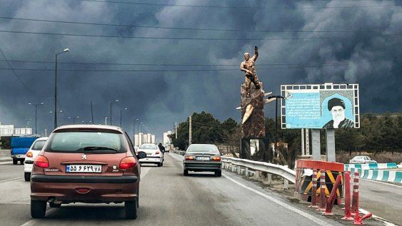 Vehículos circulan por una autopista junto a una estatua conmemorativa de guerra y una valla publicitaria que representa al difunto líder supremo iraní, el ayatolá Alí Jamenei, quien murió en un ataque aéreo el 28 de febrero, con columnas de humo negro en Teherán el 8 de marzo de 2026.
