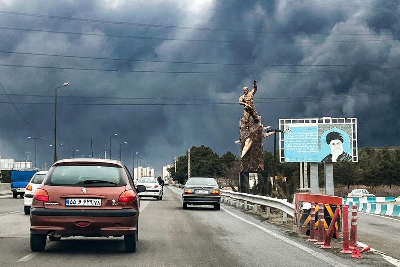 Vehículos circulan por una autopista junto a una estatua conmemorativa de guerra y una valla publicitaria que representa al difunto líder supremo iraní, el ayatolá Alí Jamenei, quien murió en un ataque aéreo el 28 de febrero, con columnas de humo negro en Teherán el 8 de marzo de 2026.
