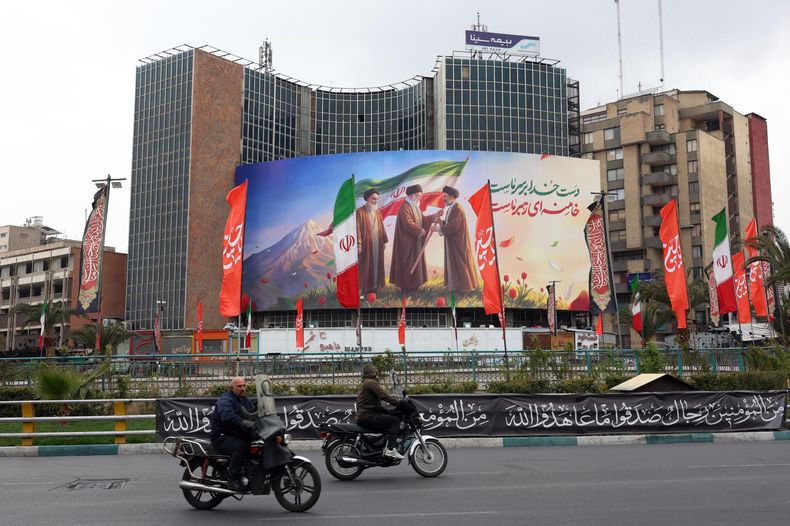 La gente pasa frente a una enorme valla publicitaria que muestra a los difuntos líderes supremos iraníes, el ayatolá Ruhollah Jomeini (izq.), el ayatolá Alí Jamenei (centro), y el nuevo líder supremo iraní, el ayatolá Mojtaba Jamenei (der.), en la plaza Valiasr de Teherán.