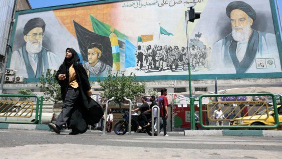Personas de origen iraní pasan junto a un cartel con imágenes de los difuntos líderes supremos iraníes, el ayatolá Ali Khamenei (arriba a la izquierda) y Ruhollah Khomeini (arriba a la derecha), en una calle de Teherán, Irán, el 18 de mayo de 2026.&nbsp;
